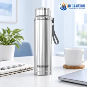 Bình Sunhouse Flexi inox có dây đeo xám đứng cạnh sổ và cốc trên bàn sáng.
