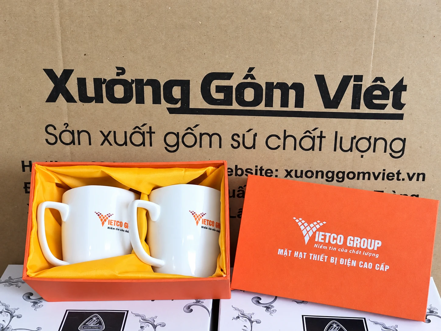 Hai cốc sứ trắng in logo VIETCO GROUP trong hộp cam lót vải vàng, nắp hộp đặt bên cạnh.