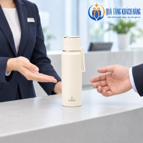 Bình Kalpen AR15 trắng kem tại quầy tiếp khách, có bàn tay minh họa trao và nhận quà.