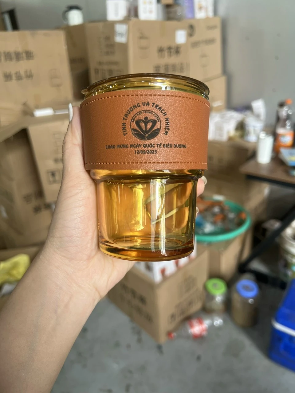 Ly thủy tinh màu hổ phách với phần bọc nâu có logo và dòng chữ đen, được cầm trên tay.