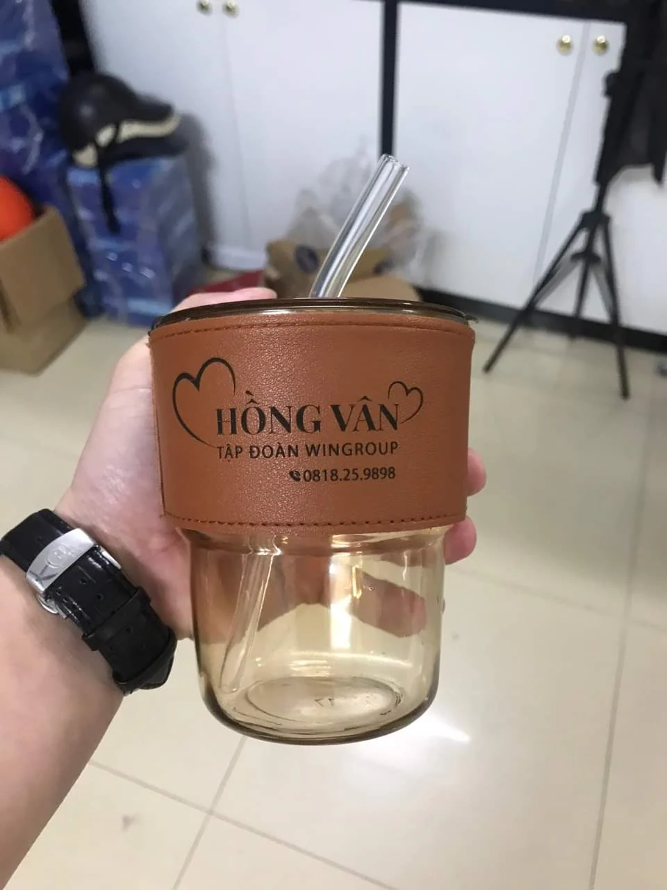 Ảnh thật ly thủy tinh có phần bọc nâu in logo Hồng Vân, nắp và ống hút trong suốt, được cầm trên tay.