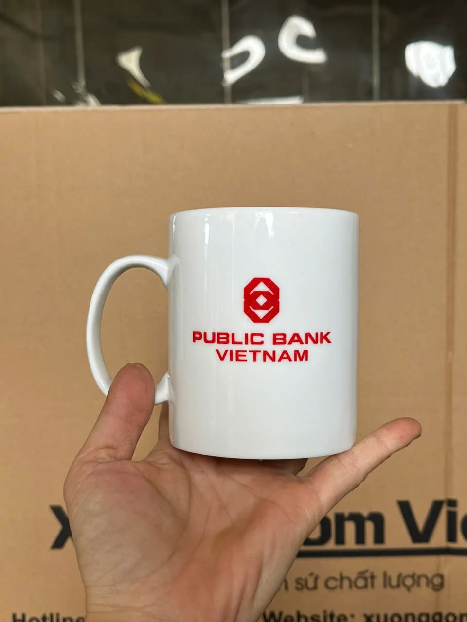 Cốc trắng mang chữ PUBLIC BANK VIETNAM được đỡ trên tay, nhìn rõ quai và vùng in trong ảnh nguồn.