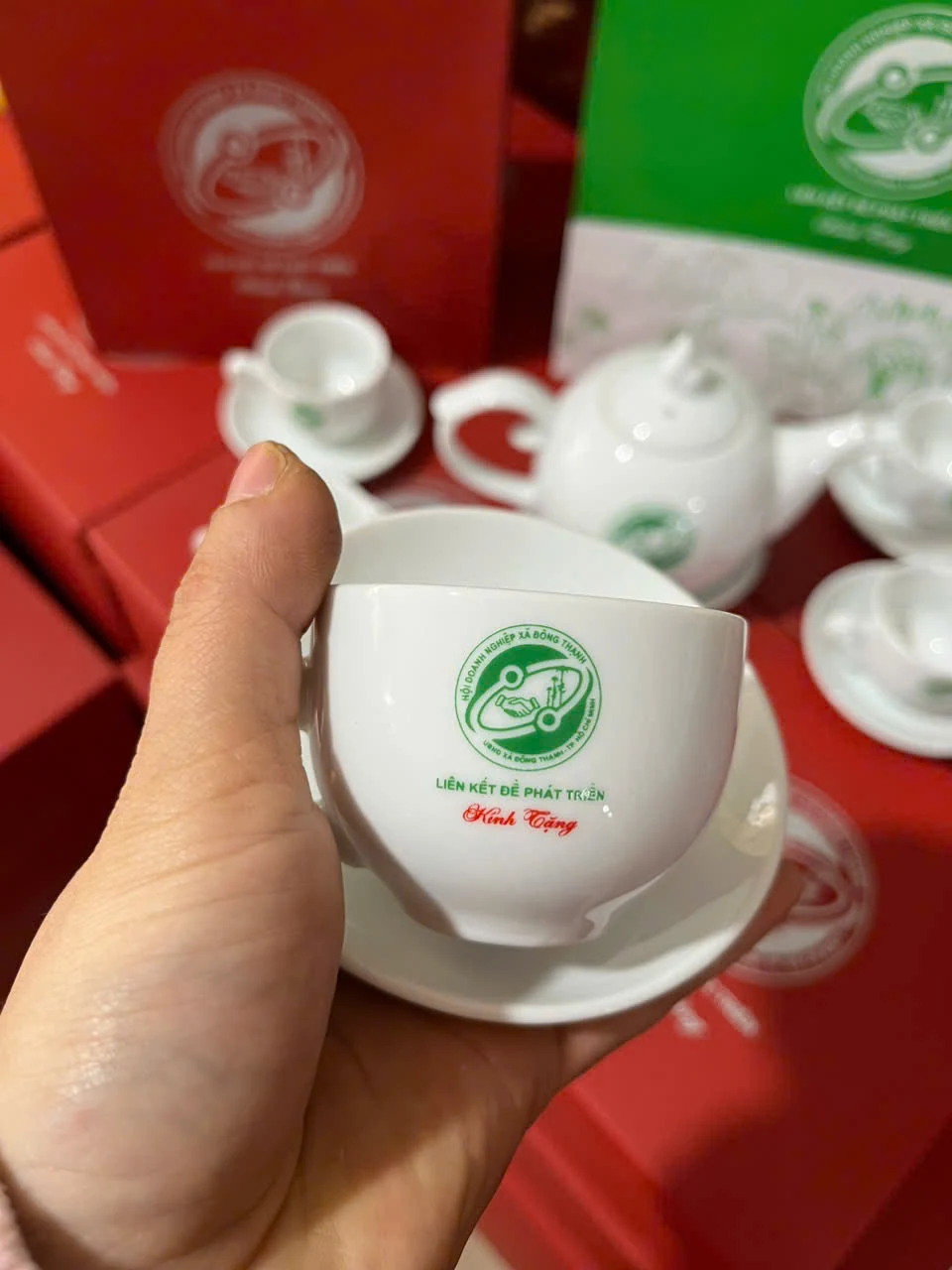Chén sứ trắng in logo xanh và chữ Kính Tặng, phía sau có ấm, chén và hộp đồng bộ.