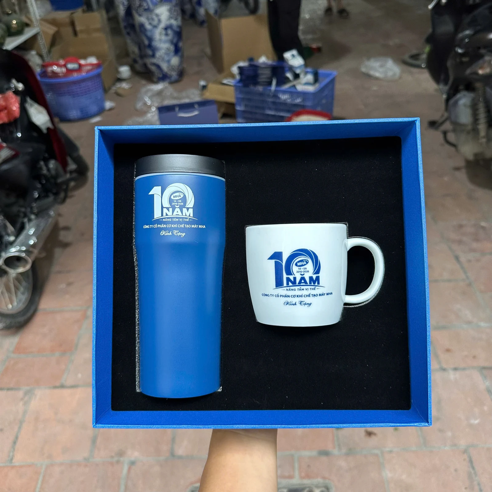 Bình xanh có nắp và cốc trắng cùng nhận diện kỷ niệm trong hộp ở ảnh thật.