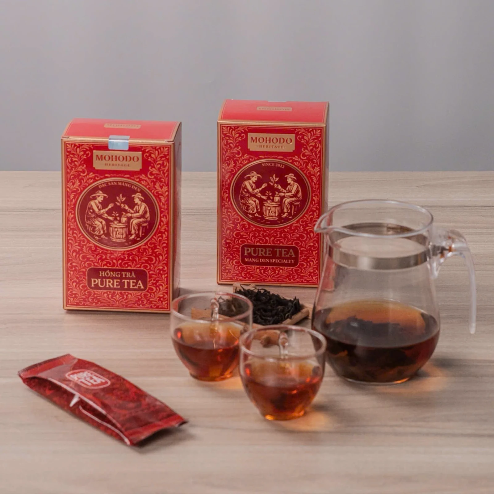 Hai hộp đỏ MOHODO Pure Tea cạnh gói trà, bình và cốc trong ảnh nguồn.