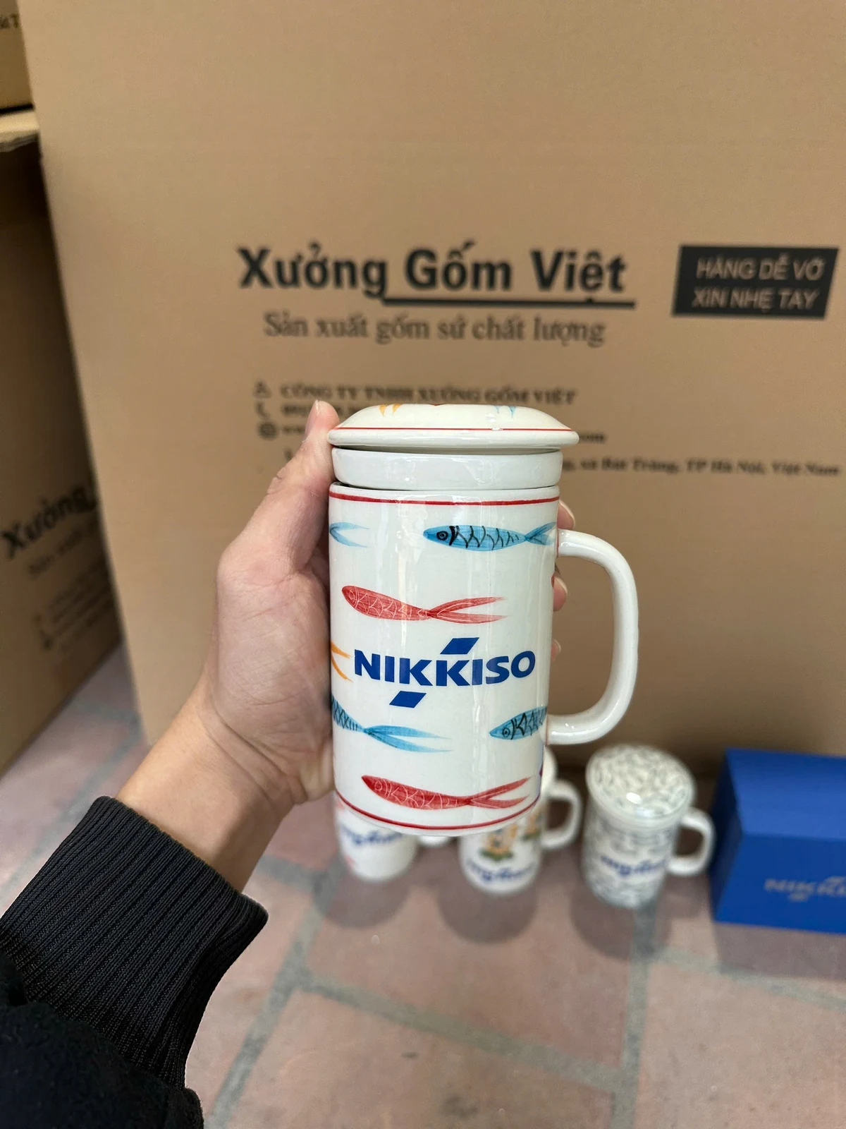 Ảnh thật tay cầm cốc trắng có quai, nắp và hoa văn cá, logo NIKKISO ở mặt trước.