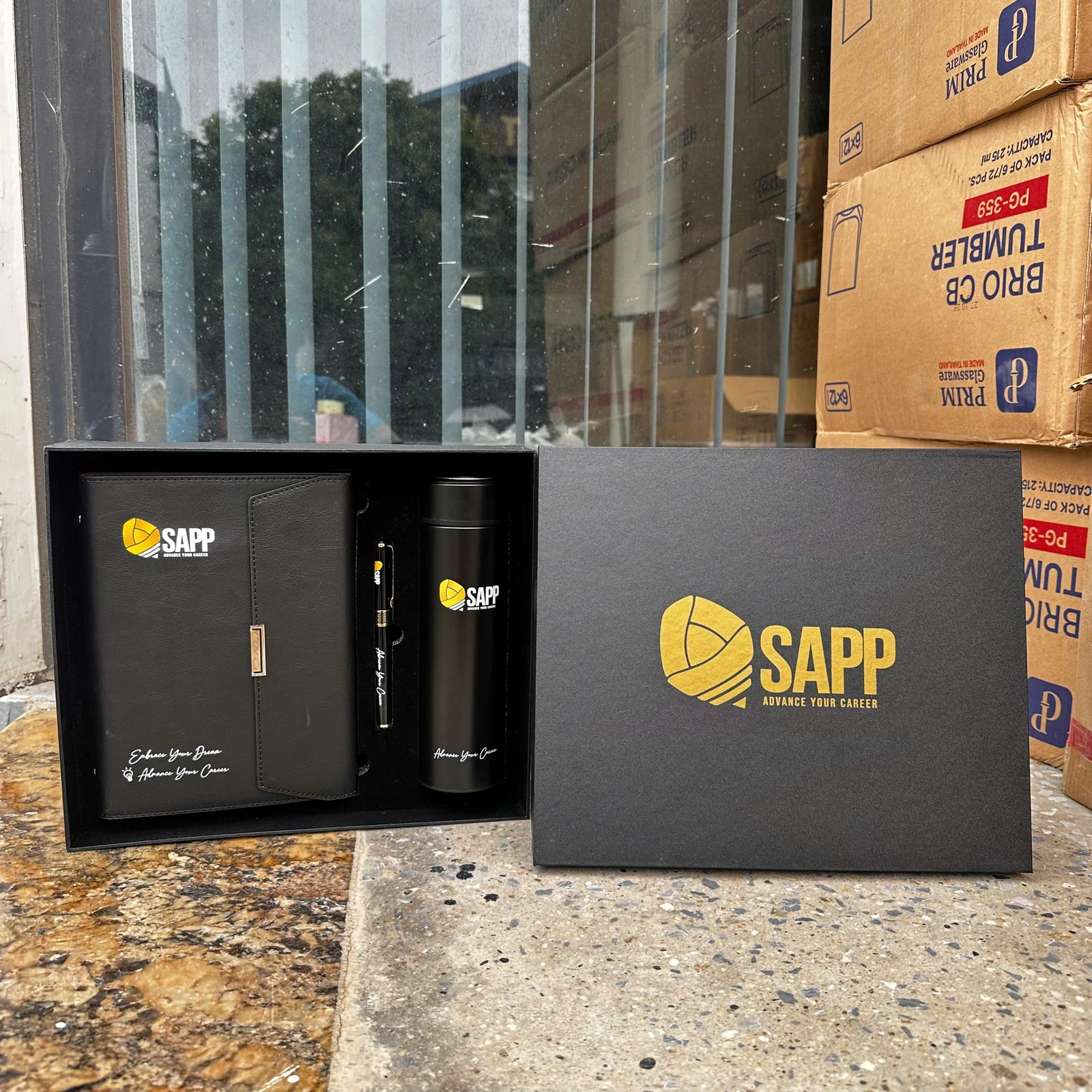Bộ quà đen có sổ, bút và vật phẩm đồ uống cùng nhận diện SAPP trong ảnh nguồn.