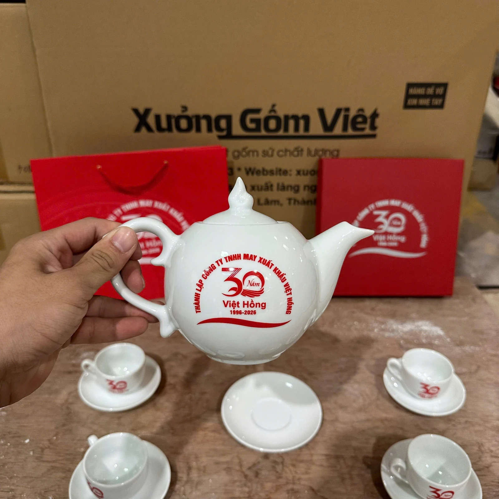 Bàn tay cầm quai ấm trà trắng có logo đỏ; vòi và nắp hiện rõ.