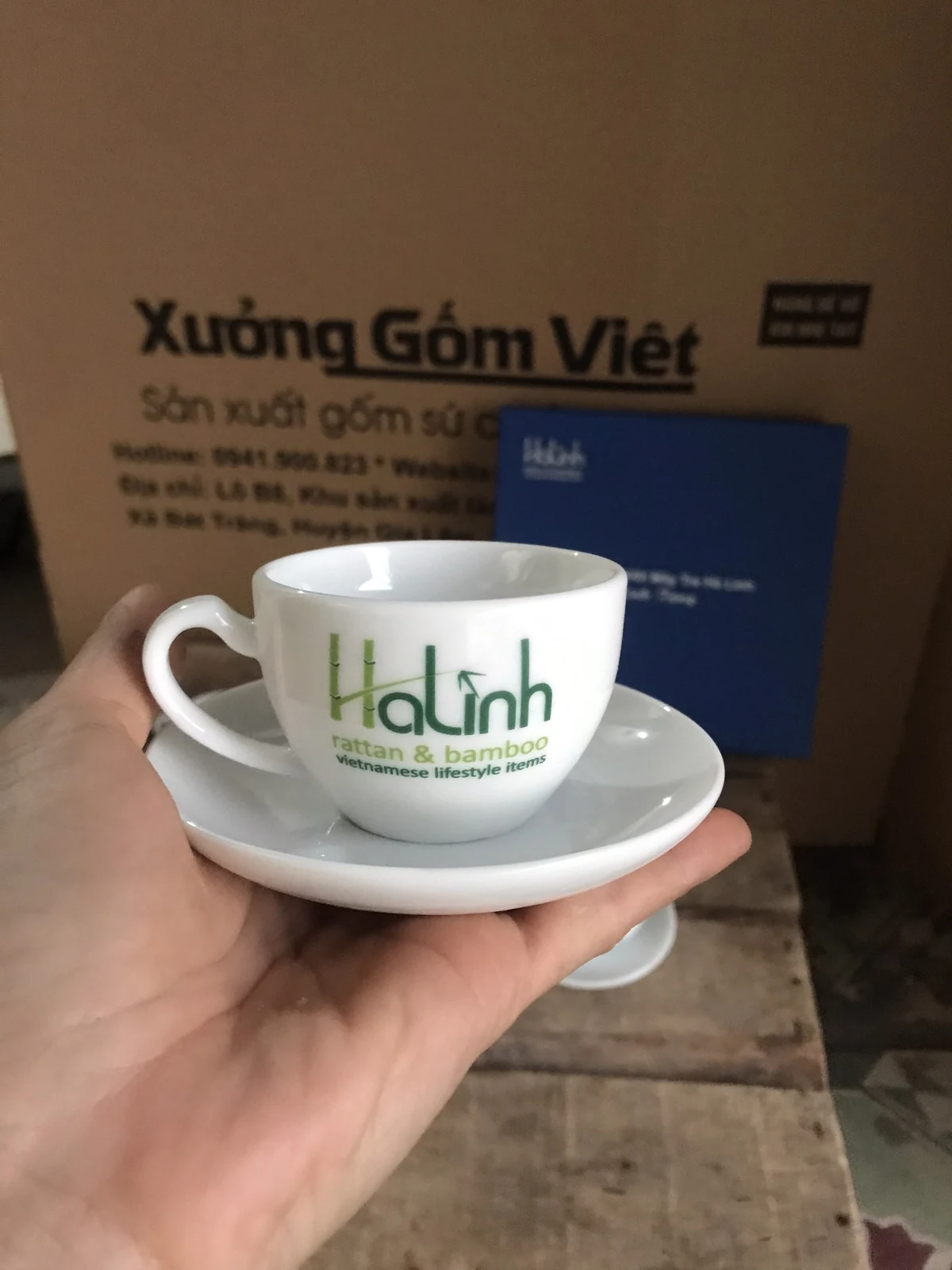 Tách trà trắng có quai và đĩa lót, mặt in logo xanh Hà Linh hướng về phía người xem.