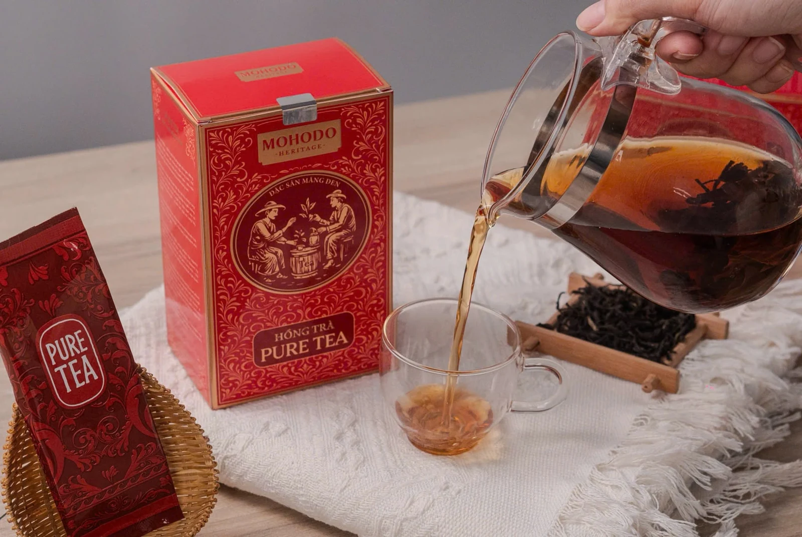Ảnh nguồn thao tác rót trà từ bình thủy tinh cạnh hộp và gói Pure Tea.