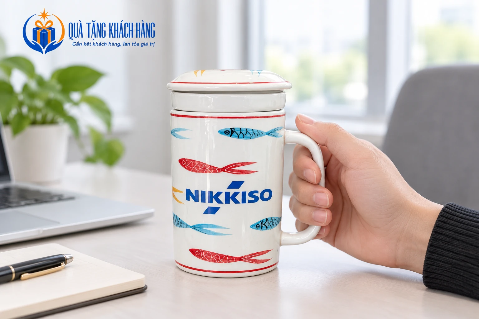 Cốc sứ có nắp, quai và họa tiết cá được cầm tại bàn, giữ logo NIKKISO của mẫu nguồn.