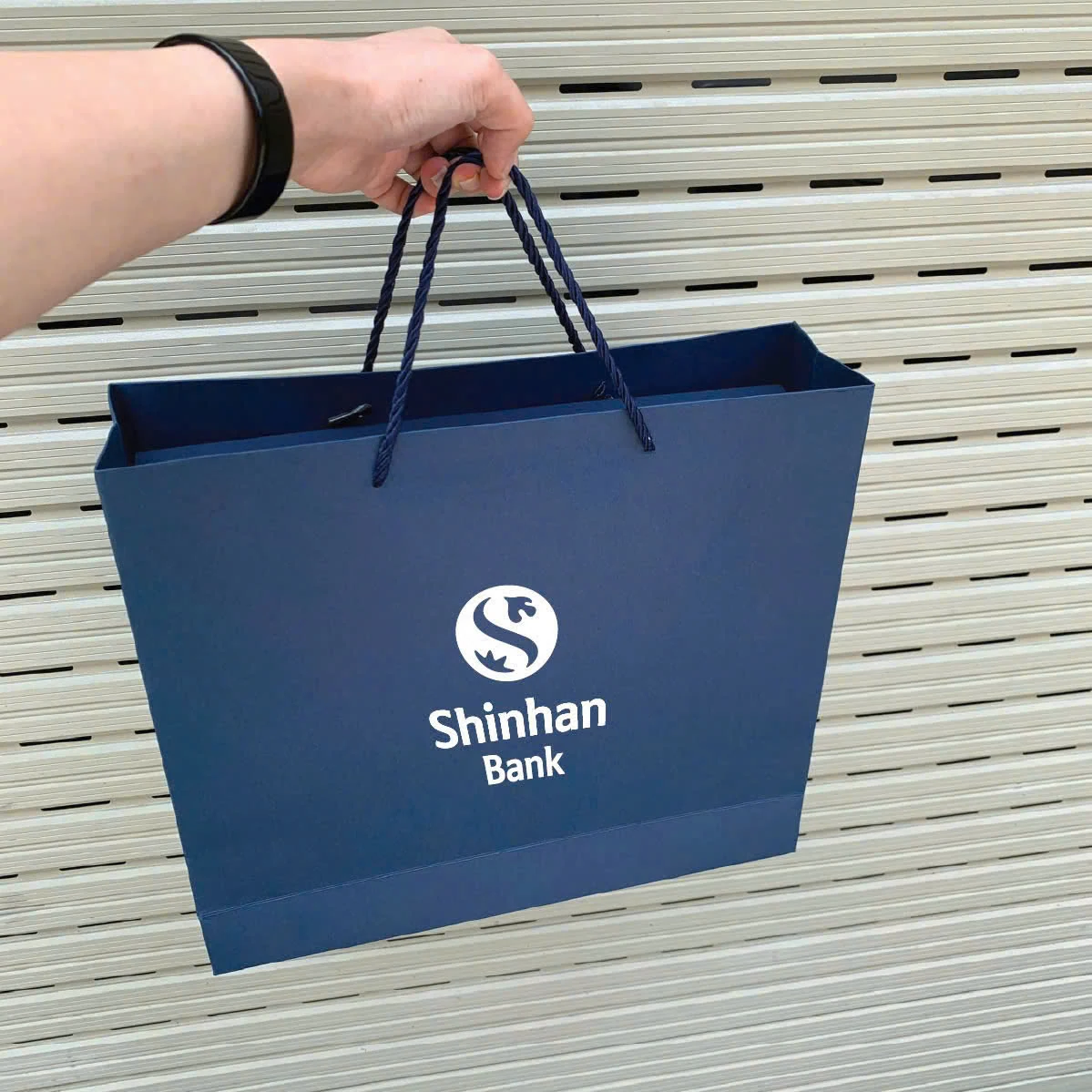Túi giấy xanh in Shinhan Bank được cầm bằng quai dây xanh.