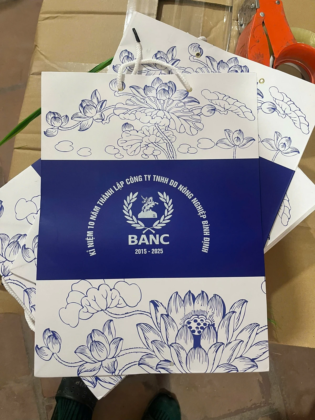 Túi giấy trắng xanh in hoa sen, tên BANC và mốc kỷ niệm 2015–2025.